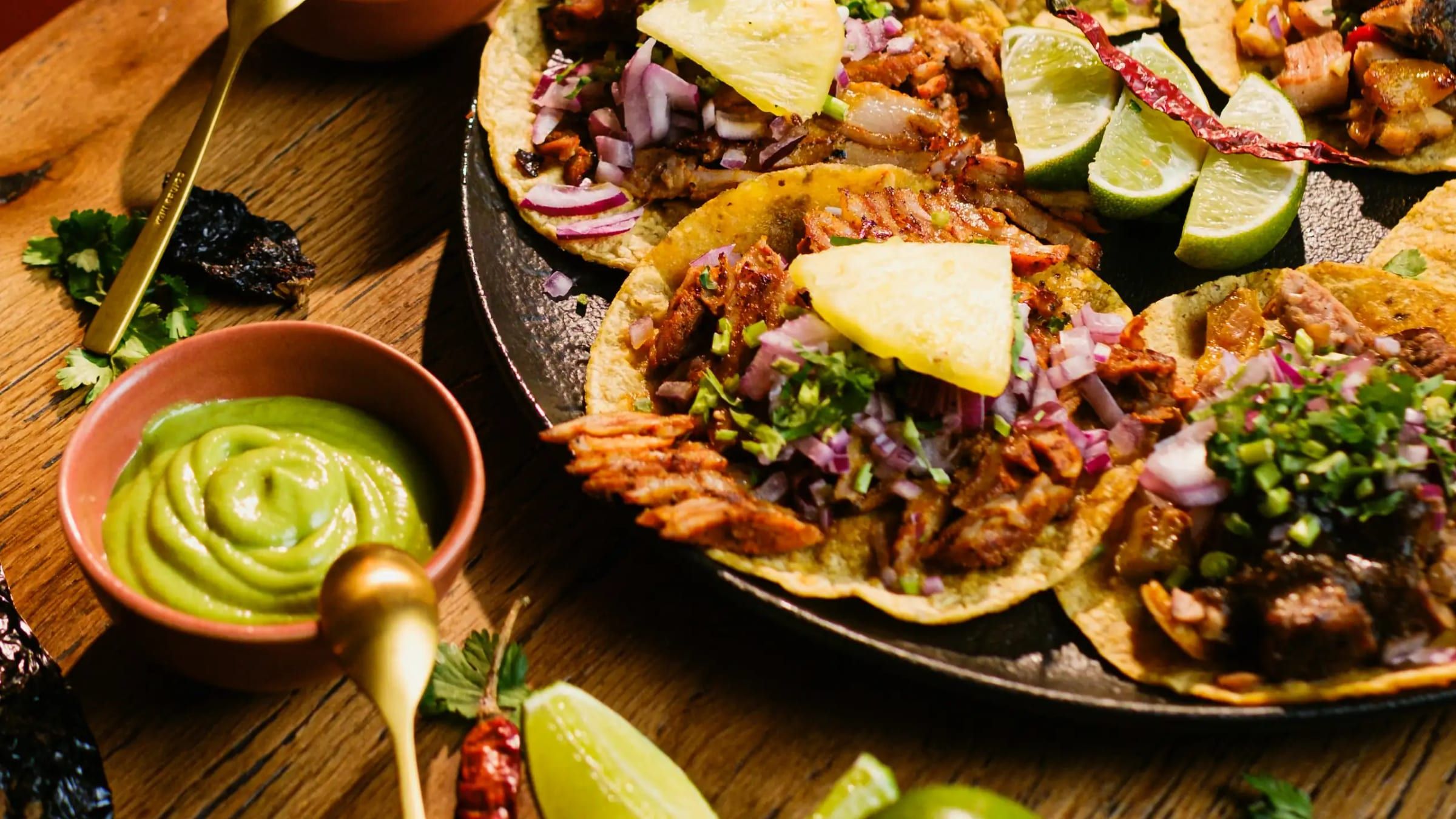 Al pastor tacos topped with pineapple, red onion and cilantro, with salsas, limes and dried chiles