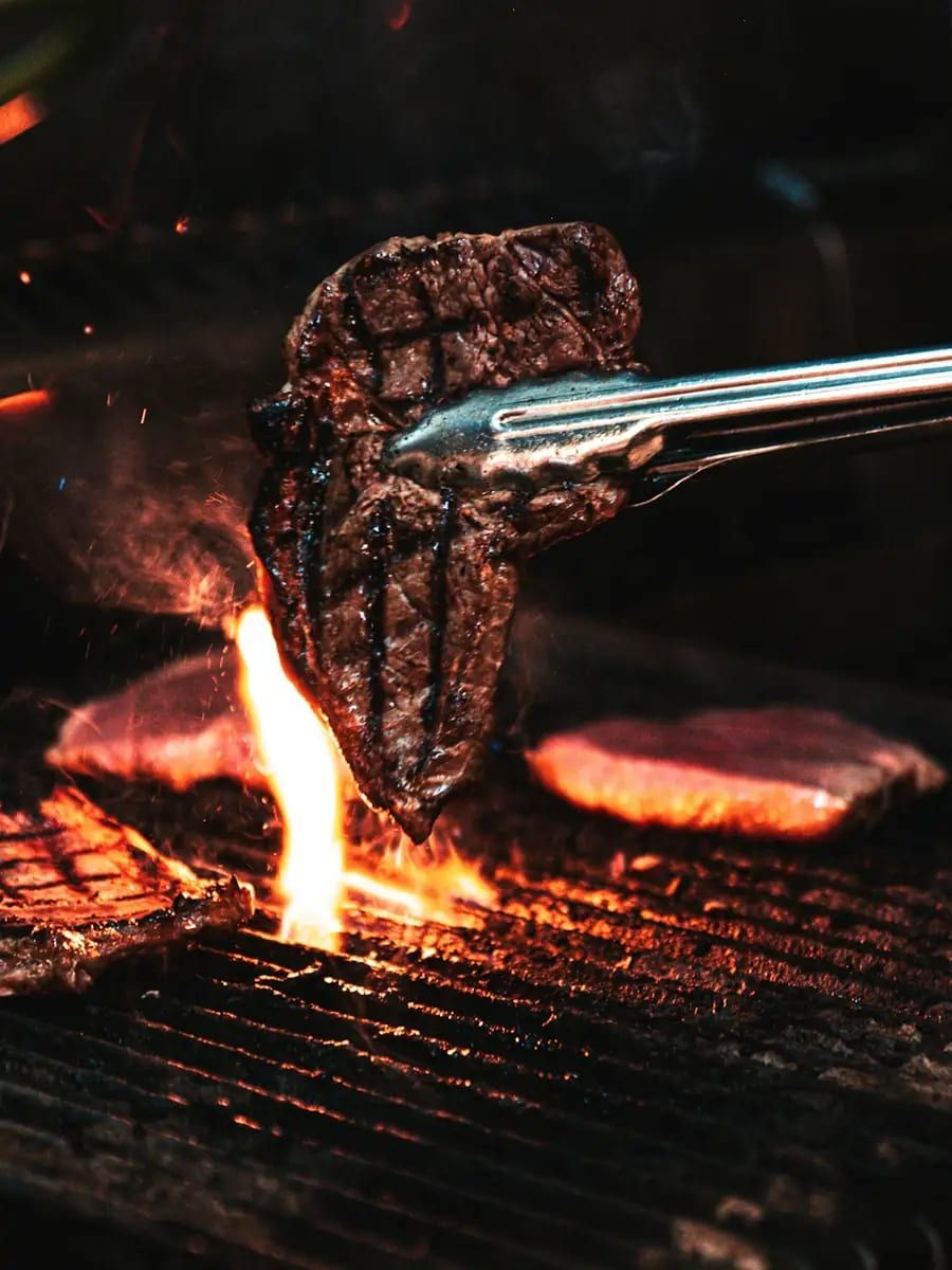 Tongs lifting a seared steak over a flaming charcoal grill