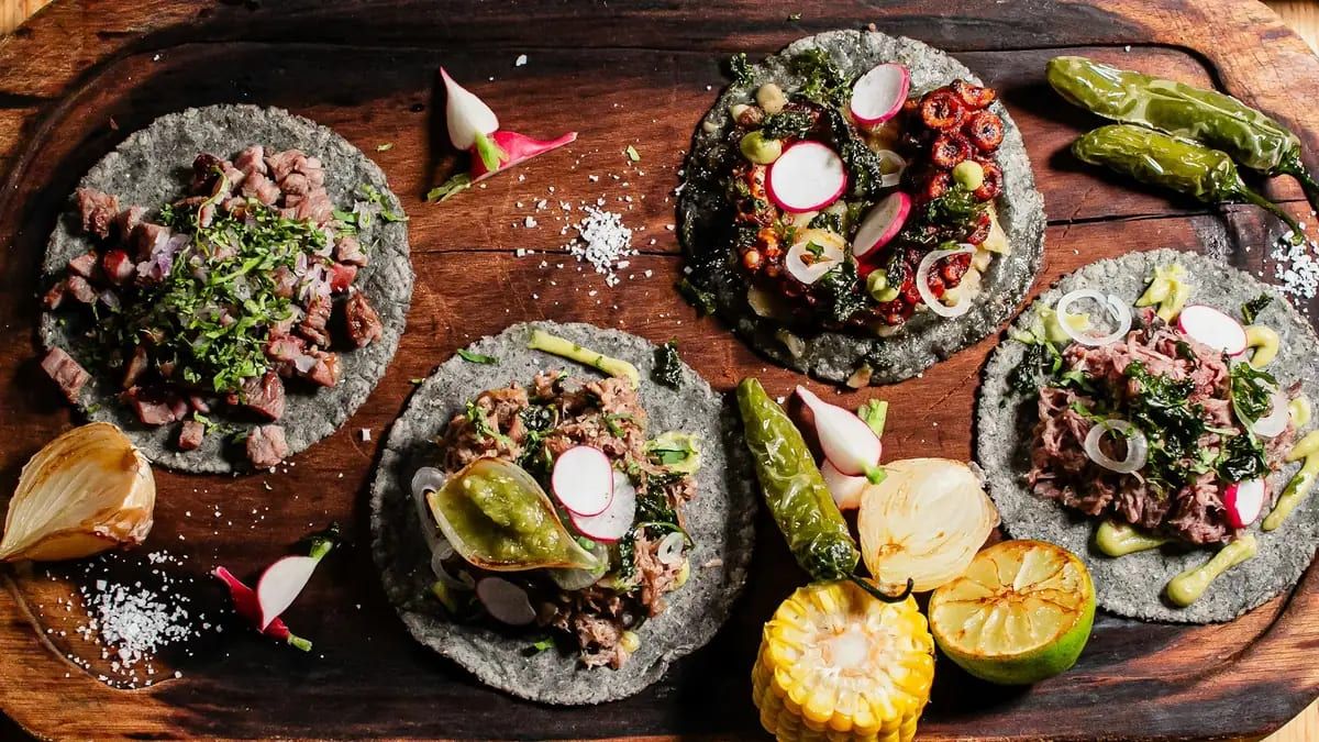 Four tacos on blue-corn tortillas on a wooden board with charred onion and lime